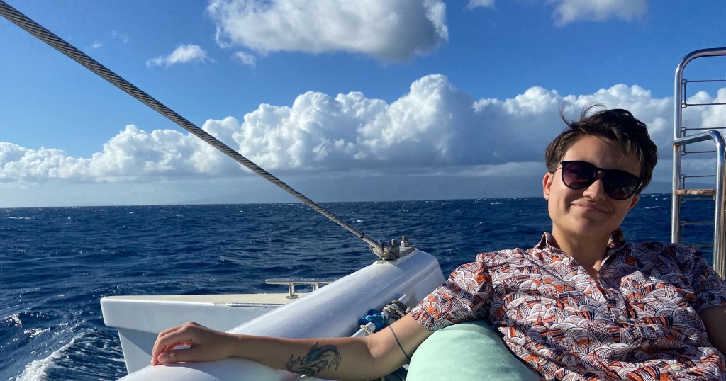 Calenn laying back in the bay of a boat on a dark blue sea. Water surges beneath the boat and big fluffy white clouds are visible on the horizon.